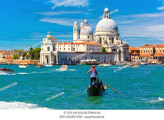 Gondolier in a gondola rides on Canal Grande in a hat with a red ribbon and a typical striped singlet, Basilica di Santa Maria della Salute in the background