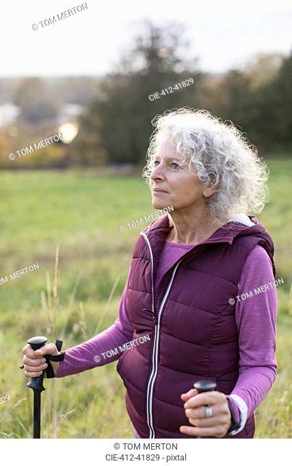 Thoughtful active senior woman hiking with poles in rural field