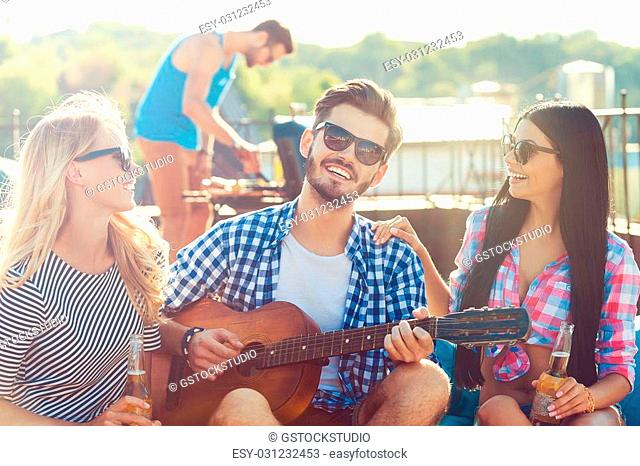 Sharing good time. Three cheerful young people bonding to each other and sitting on the bean bag with guitar while man barbecuing in the background