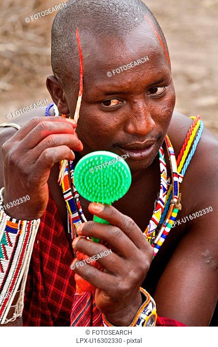 Masai applying paints for celebrations: Masai warrior dressed in traditional red shuka and much beaded jewellery sits applying red paint to his face with reed