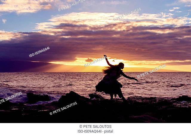 Silhouetted woman hula dancing on coastal rocks wearing traditional costume at sunset, Maui, Hawaii, USA