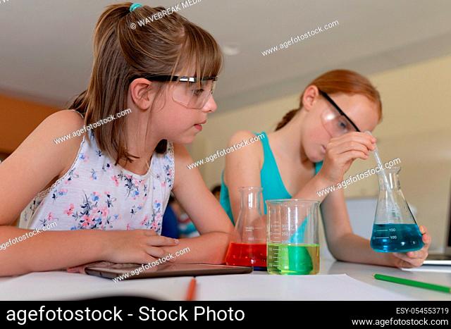 Front view close up of two Caucasian schoolgirls sitting at a desk wearing safety glasses and using flasks of colourful liquid and a pipette while doing an...