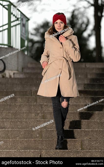 Smiling pretty young woman with long brunette hair, a light brown cloth coat and a red woolen cap walks down a staircase of natural stones