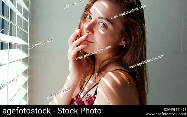 Portrait of beautiful red hair girl in lingerie posing near the window with blinds during sunny day morning
