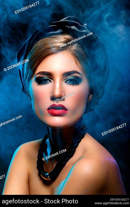 Beautiful young woman with bright makeup, red lips, black shawl on head and necklace. Studio beauty shot with blue smoke. Copy space
