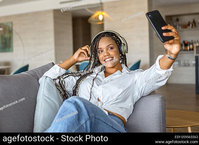 Front view of a happy young woman listening to music with headphones and using mobile phone to take a selfie while leaning on a sofa at home