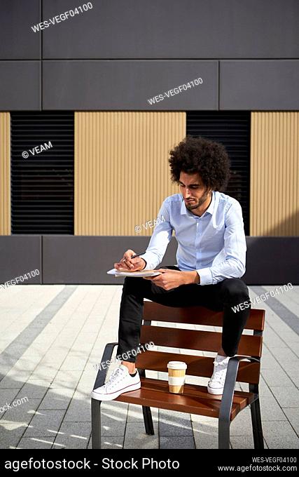 Man with coffee cup writing in book while sitting on chair during sunny day