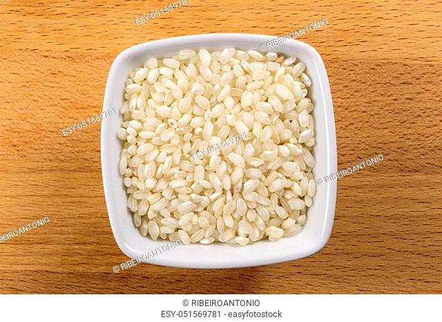 Overhead shot of raw arborio short grain rice in a white bowl on a wooden chopping board background. Arborio rice is the main ingredient of the traditional...