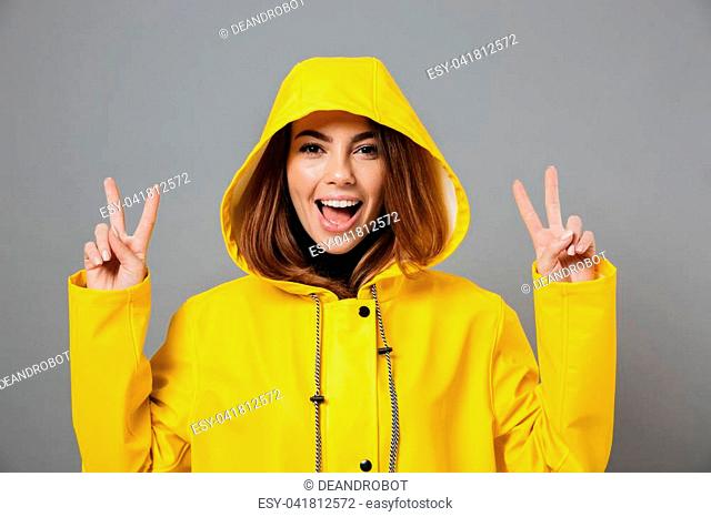 Close up portrait of a cheery girl dressed in raincoat posing with hood on her head and showing peace gesture isolated over gray background