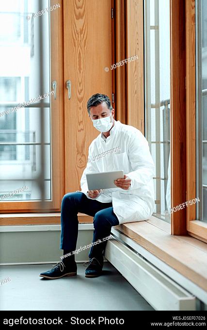 Scientist with protective face mask using tablet PC sitting on window sill at medical clinic
