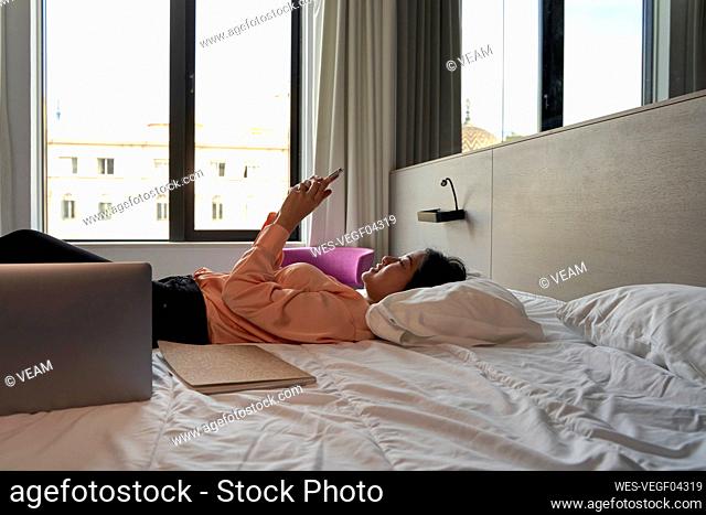 Female professional text messaging through smart phone while lying on bed in hotel
