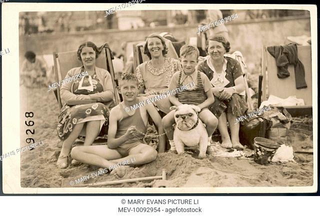 On the beach at Margate: three ladies sit in deckchairs, while two boys in bathing costumes sit, one on the sand, one astride a toy bulldog