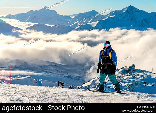 A professional snowboarder with a backpack stands on the snow high in the mountains against the background of low clouds and the Caucasus mountains during a...