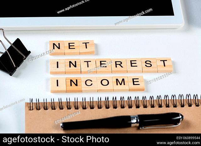 Net interest income concept with letters on wooden cubes. Still life of office workplace with supplies. Flat lay white surface with tablet computer and notepad