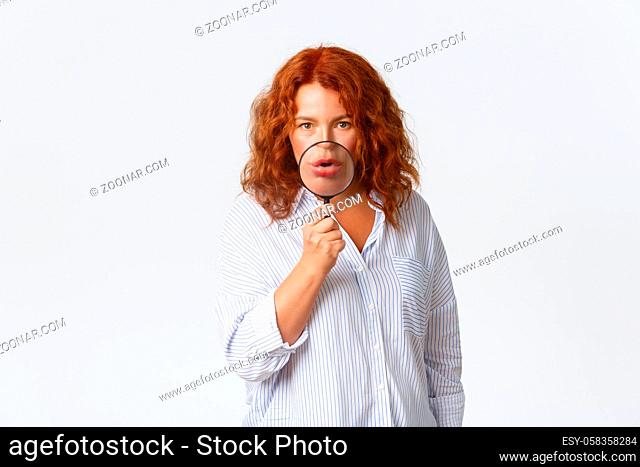 Portrait of redhead middle-aged woman searching for something, holding magnifying glass over mouth and looking funny at camera