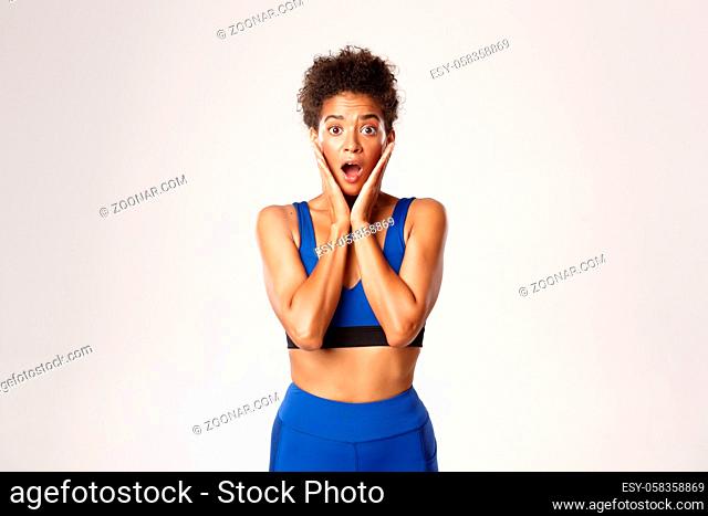 Waist-up of surprised african-american fit female athelte, looking amazed, open mouth wondered, standing against white background in blue sport outfit