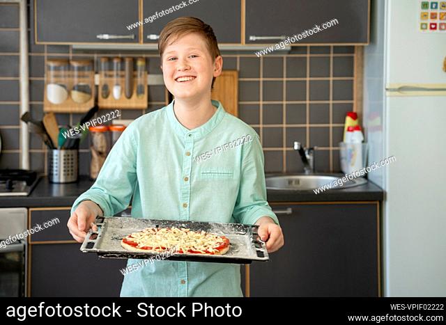 Portrait of proud boy holding baking tray with raw homemade pizza
