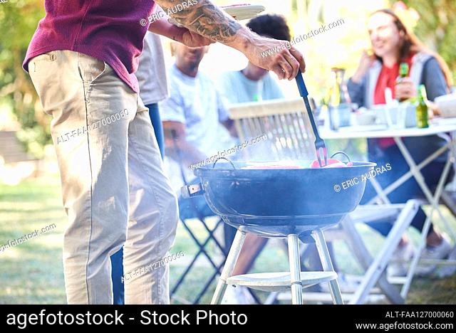Young man preparing food in barbecue while his friends sitting in background