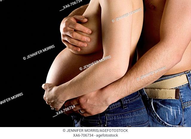 A pregnant woman wearing jeans and nothing else, is cradled from behind by her husband, similarly attired. No faces are shown. Black background