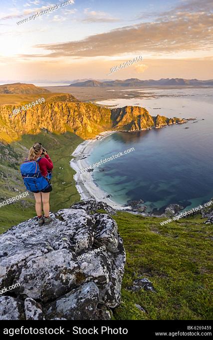Hiker standing on a rocky outcrop overlooking beach and sea, summit of Måtinden mountain, near Stave, Nordland, Norway, Europe