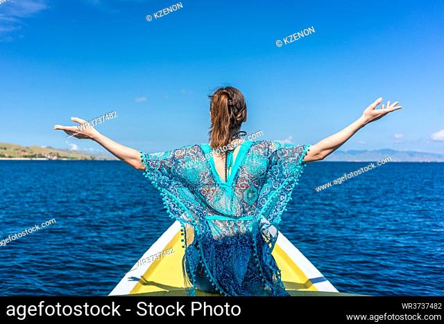 Rear view of a young woman sitting with outstretched arms against a clear blue sky while enjoying summer vacation in Flores Island, Indonesia