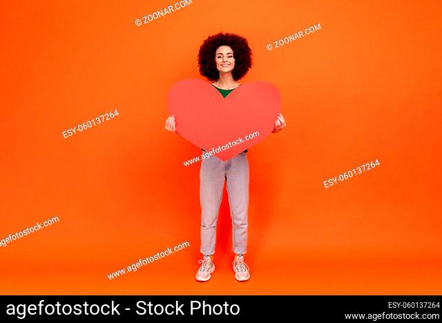 Full length portrait of falling in love woman with Afro hairstyle wearing green casual style sweater holding big red heart, looking with toothy smile