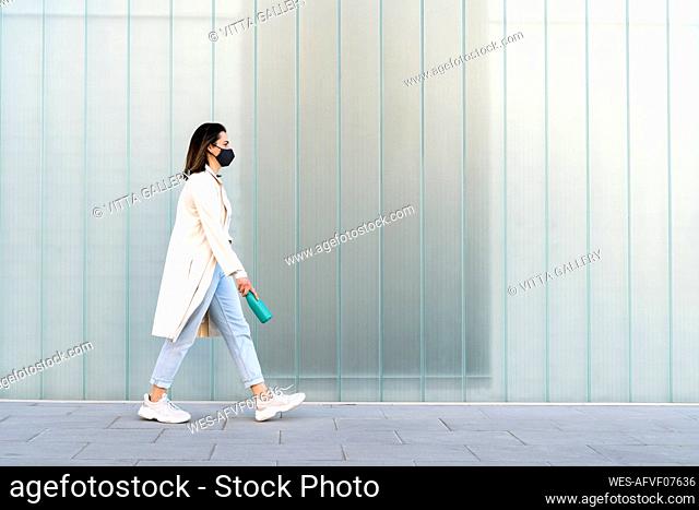 Businesswoman with bottle walking on footpath by glass wall during pandemic