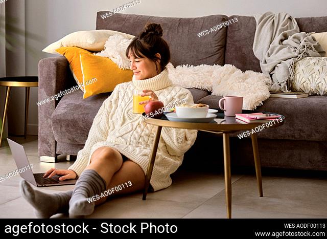 Woman in sweater having juice while using laptop at home