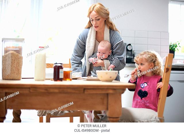 Mother holding baby boy, sitting at kitchen table with young daughter, having breakfast