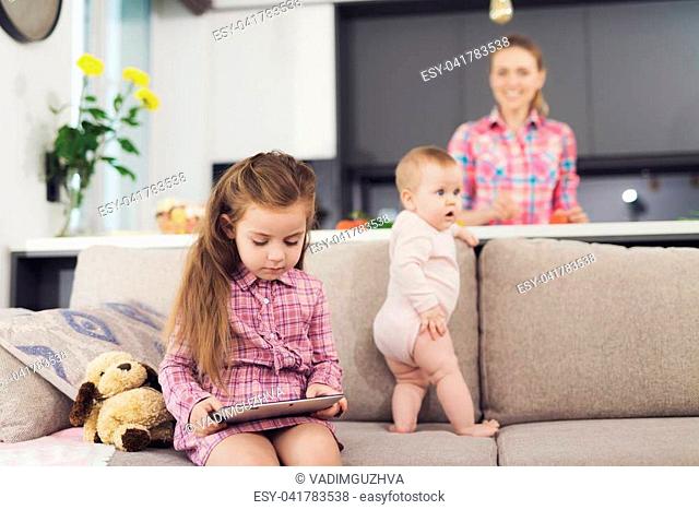 A girl and a baby are sitting on the couch. The girl is looking at something on the tablet while her sister is holding onto the pillow and looking away