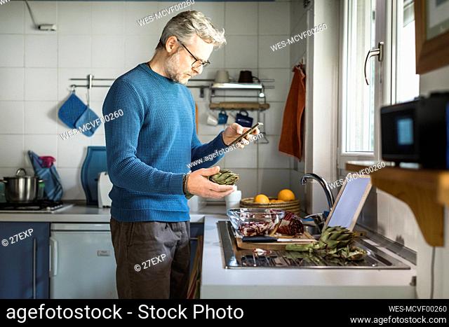 Mature man preparing artichoke in his kitchen looking at smartphone