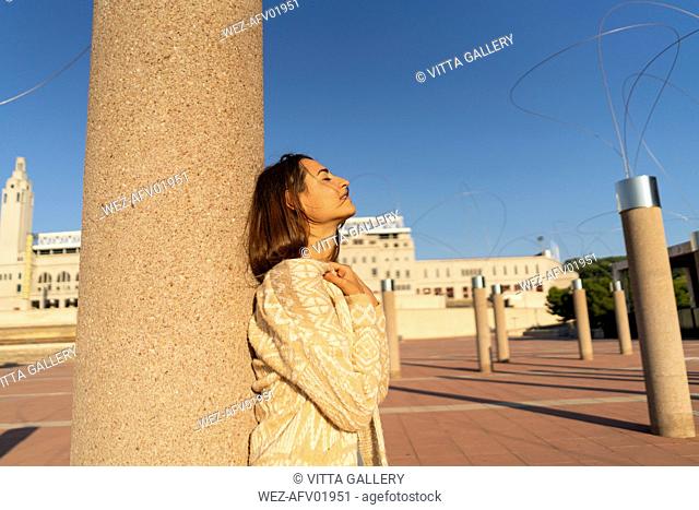 Spain, Barcelona, Montjuic, young woman leaning against a column in sunlight