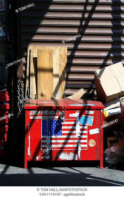 A red garbage dumpster with American flag stands on pavement covered with shadows cast by staircase
