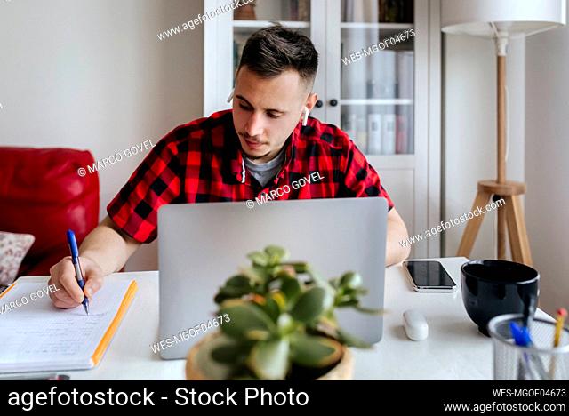 Handsome male entrepreneur with in-ear headphones writing while working on laptop at home office