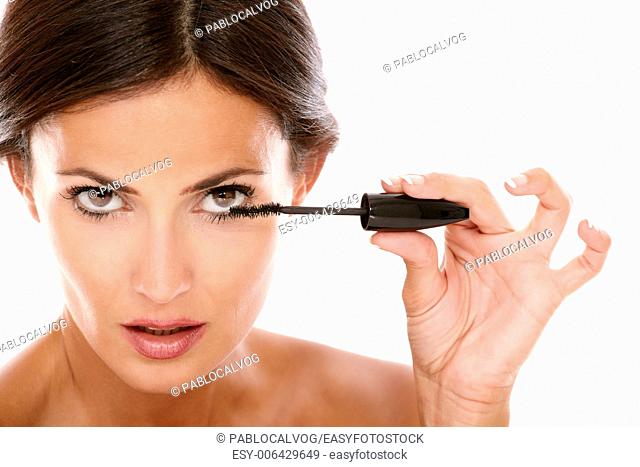 Front view portrait of young latin woman using mascara to her eyelash with nude shoulders while looking at camera on isolated studio