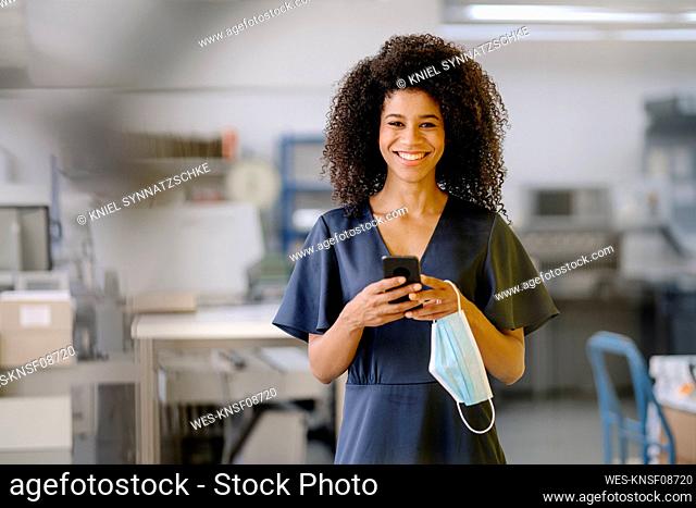 Smiling businesswoman with mobile phone and protective face mask standing in office