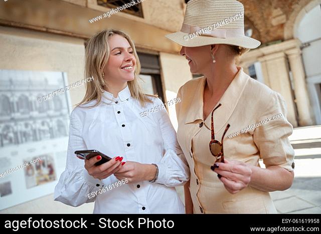 Pleased blonde young woman with the smartphone looking at a smiling stylish lady with sunglasses