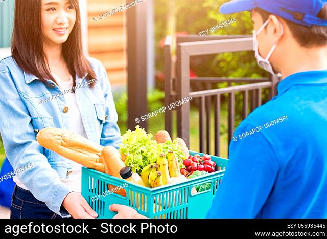 Asian young delivery man wear face mask making grocery fast service giving fresh food vegetable in plastic basket to woman customer receive at house door after...