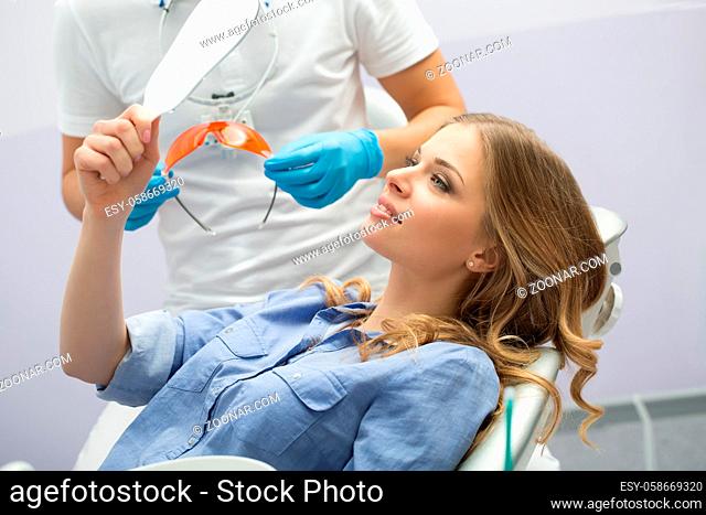 Pretty girl in blue shirt with a mirror in the right hand on the patient chair. Next to her there is a dentist in a white uniform with blue latex gloves and...