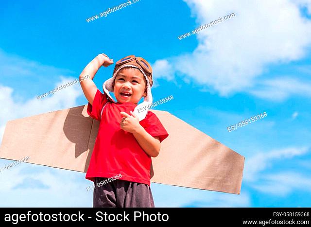 Happy Asian funny child or kid little boy smile wear pilot hat and goggles play toy cardboard airplane wing flying raises hand up against summer blue sky cloud...