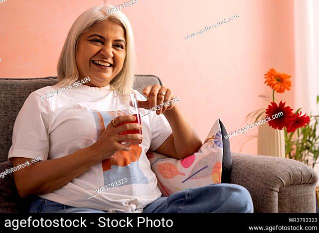 Cheerful old woman drinking soft drink while sitting on sofa at home