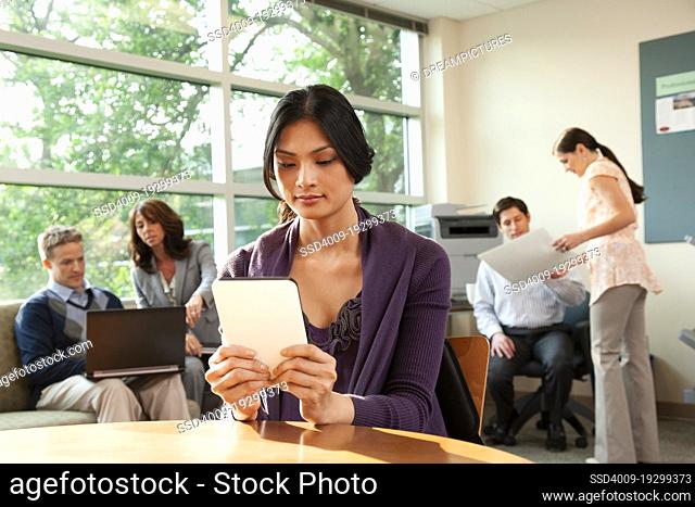 Woman sitting at table in breakroom working on tablet with coworkers behind her