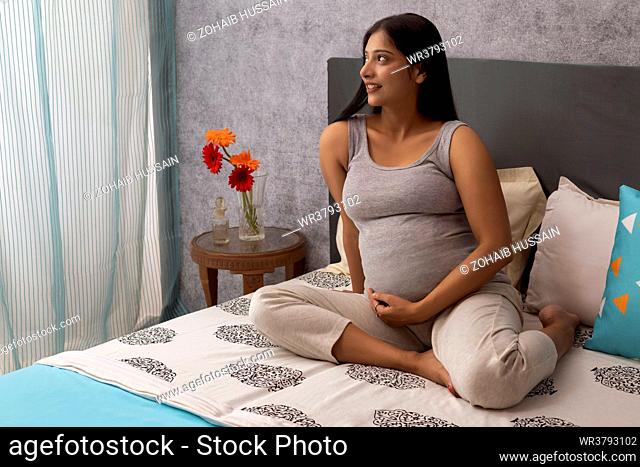 Pregnant woman looking out of window while sitting on bed at home