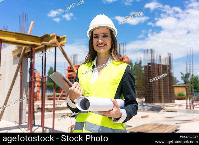 Portrait of a confident female architect or engineer with can-do attitude smiling while holding a rolled blueprint and a tablet on the construction site