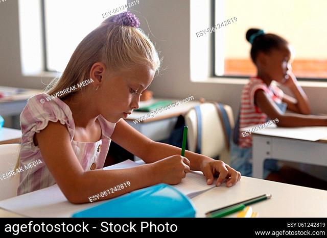 Caucasian elementary schoolgirl writing while studying at desk in classroom
