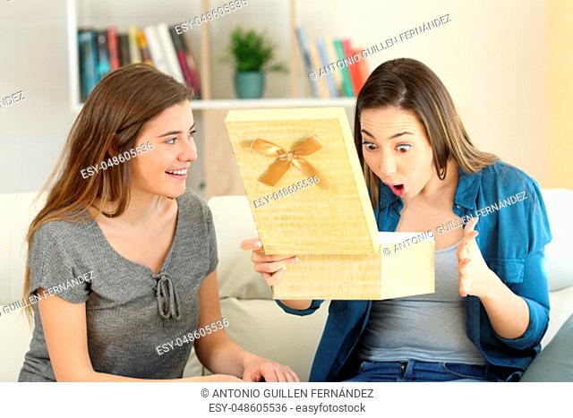 Woman giving a gift to her amazed friend sitting on a couch in the living room at home