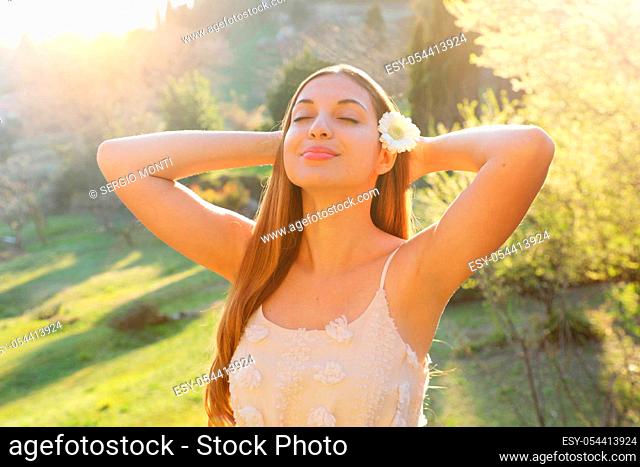Relax in nature. Beautiful young woman with flower on ear deep breathing with closed eyes staying outdoor with crossed arms behind the head in spring on sunset