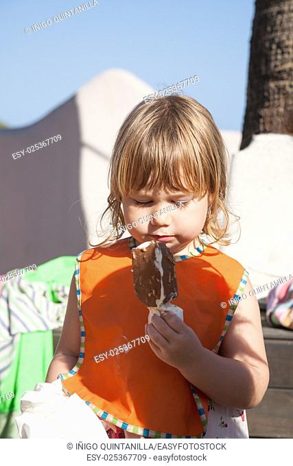 portrait of three years old blonde happy child with orange bib eating chocolate and white popsicle ice cream in exterior