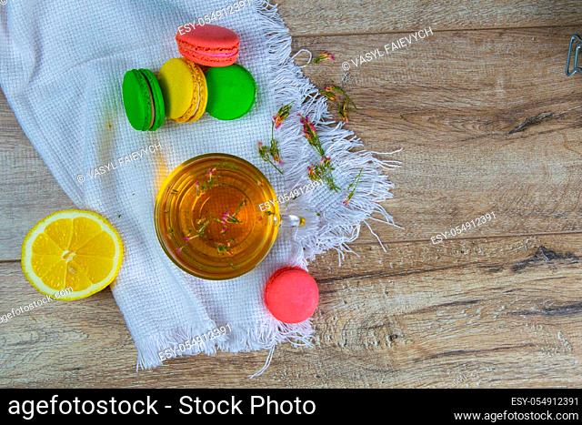 Glass transparent cup with mint tea stands on a wooden stand, next to a sugar bowl with sugar cubes, sliced lemon, spoon and multi-colored muffins