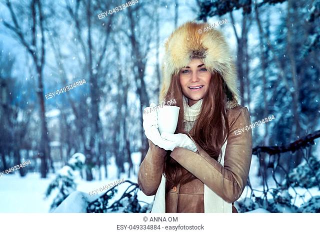 Portrait of a pretty woman wearing stylish furry hat with hot cup of tea in hands standing in the snowy park, enjoying winter holidays